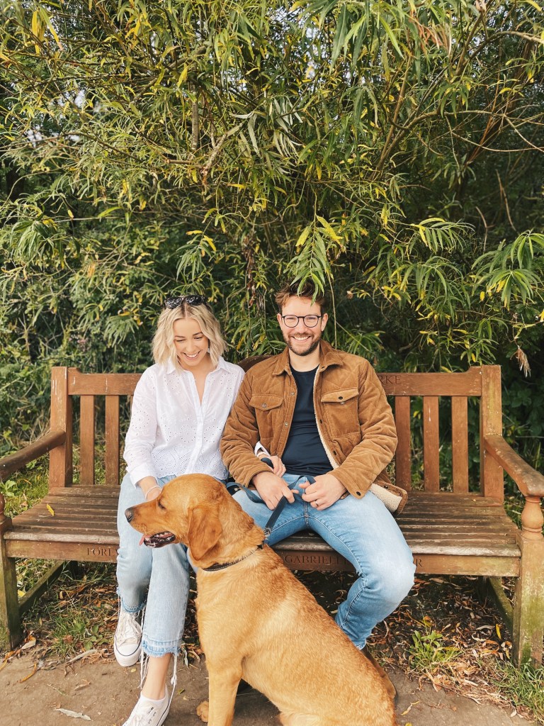 A man with glasses and a woman in a white shirt smile while sitting on a wooden bench with their dog.