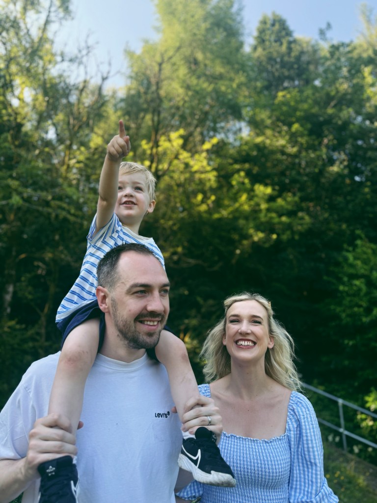 A family smiling outdoors as a young boy sits on a man's shoulders and points at the sky.