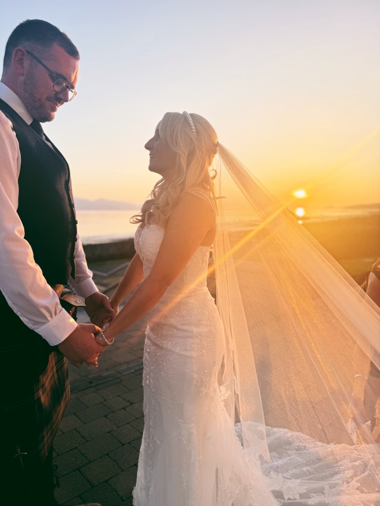 A newlywed couple standing together by the water at sunset, with golden light filtering through the bride's veil.