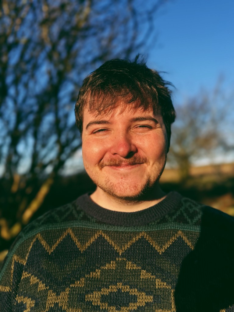 A man in a patterned, knit sweater smiling outdoors against a backdrop of bare winter trees and a clear blue sky.