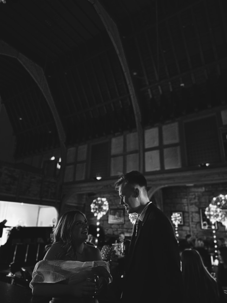 A candid black and white shot of a couple talking indoors under a high ceiling with decorative lights glowing in the background.
